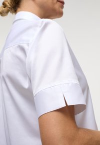 Camicia bianca a maniche corte con colletto a punta, caratterizzata da una silhouette pulita e da una fessura laterale sulle maniche per maggiore comodità.