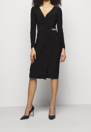 Vrouw draagt een zwart wikkeljurk tot op de knie met lange mouwen, V-hals, een decoratieve broche ter hoogte van de taille en zwarte hoge hakken.