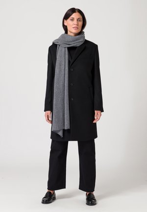 Cappotto di lana nera con chiusura a bottoni, sciarpa grigia, pantaloni neri a gamba larga e mocassini neri lucidi. Design semplice senza motivi visibili.