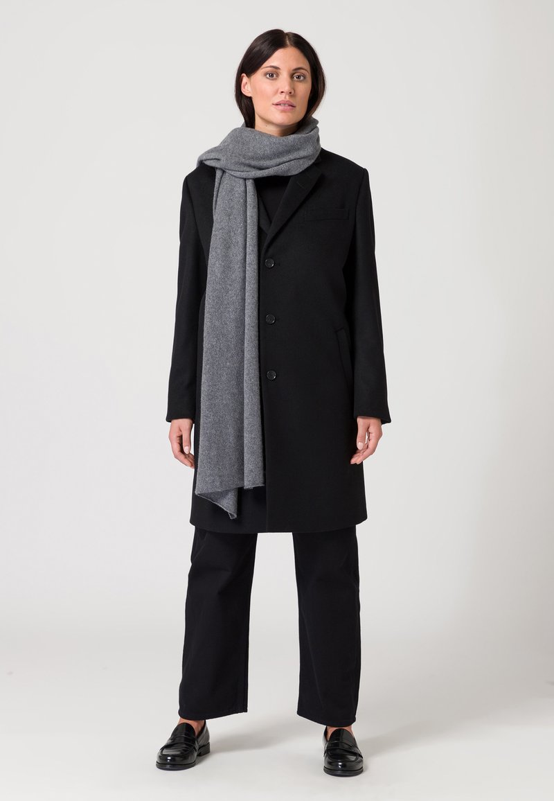 Czarny wełniany płaszcz z zapięciem na guziki, szary szalik, czarne spodnie o szerokich nogawkach i błyszczące czarne loafersy. Prosty design bez widocznych wzorów.