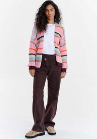 Žena s kudrnatými vlasy, oblečená v pastelově pruhovaném kardiganu, bílém tričku, tmavě hnědých kalhotách a béžových turistických botách, stojící na bílém pozadí.