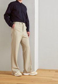 Muži v bežových kostkovaných kalhotách, bežových teniskách a tmavě námořnickém dlouhém tričku, stojící na dřevěné podlaze u světle šedé zdi.