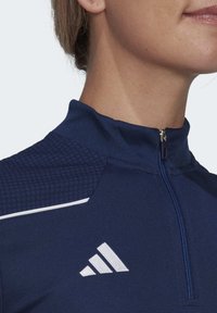 Marineblaues, zip-up Sportoberteil mit einem strukturierten Muster auf den Schultern, weißem Akzentstreifen und kleinem Logo auf der Brust. Glatte Stofftextur.