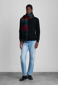 Sciarpa multicolore a righe in morbida lana, appoggiata su un maglione nero. Abbinata a jeans azzurri chiari e scarpe nere.