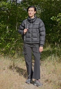 Jachetă puffer neagră, cu fermoar în față, pantaloni cargo gri și încălțăminte de drumeție. Stând pe un traseu cu copaci și iarbă în fundal.