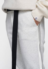 Graue Jogginghose mit schwarzem Kordelzug, ausgestattet mit einem strukturierten Muster und einem weißen gestickten Logo auf der Tasche. Weicher Stoff mit seitlichem Besatz.