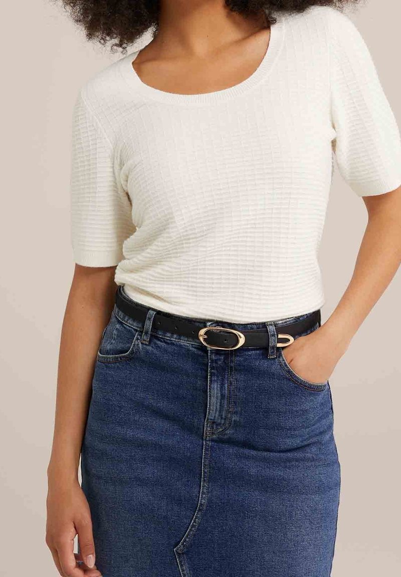 Femme portant un haut blanc texturé à manches courtes, rentré dans une jupe en denim bleue avec une ceinture noire à boucle dorée, main dans la poche.
