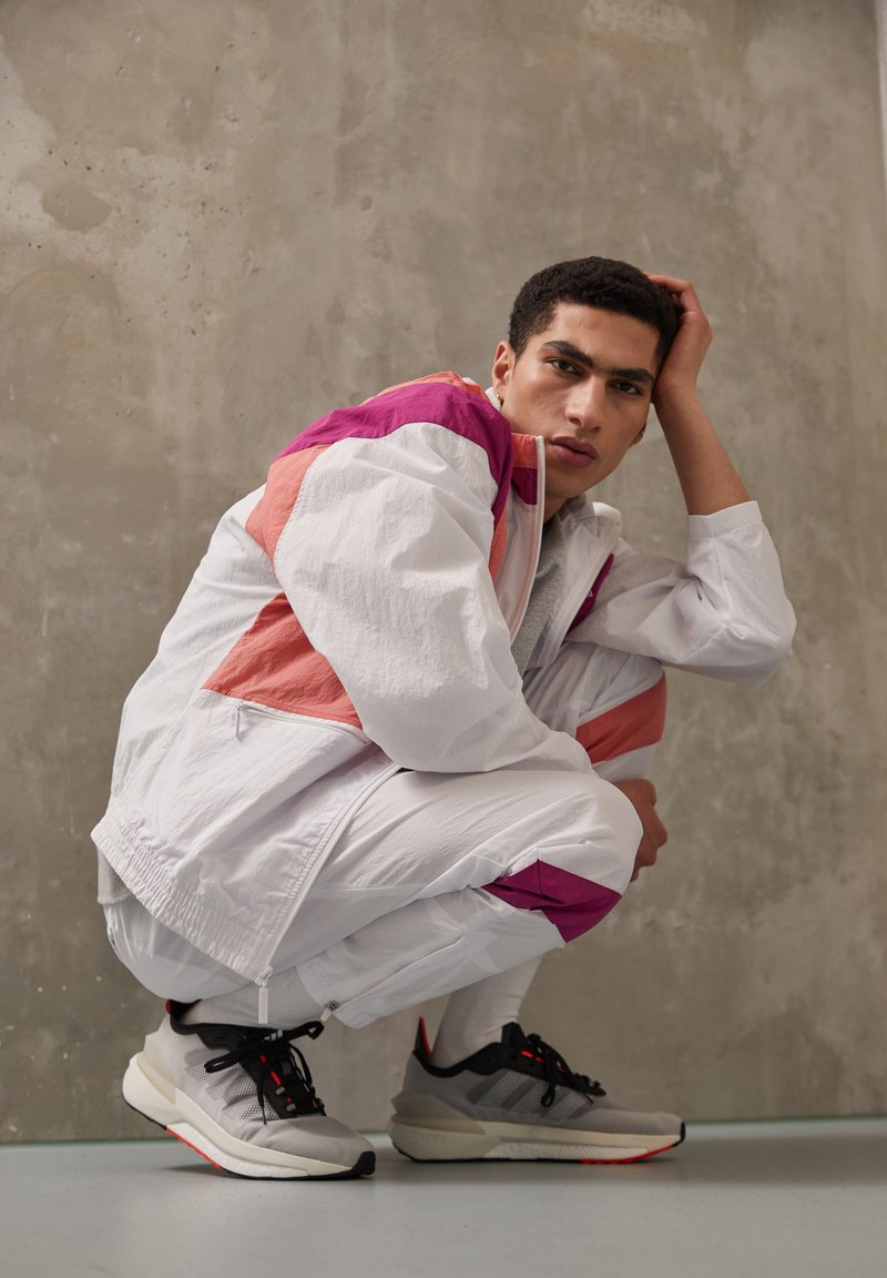 Weiße Sportjacke mit rosa und lila Akzenten, kombiniert mit grauen und schwarzen Sneakers. Modell hockend vor einem strukturierten grauen Hintergrund.