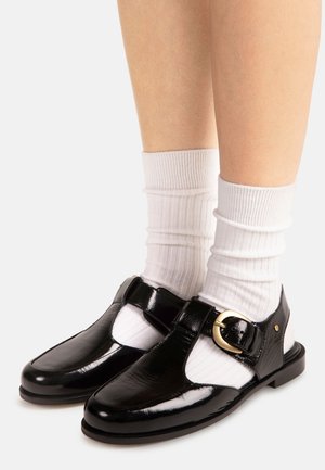 Chaussures à bride en T en cuir verni noir avec boucle dorée portées avec des chaussettes blanches côtelées sur des jambes nues