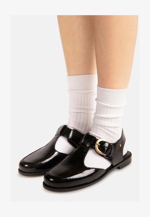 Zwarte lakleren T-strap schoenen met gouden gesp, gedragen met witte geribbelde sokken tot aan de kuit op blote benen