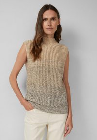Frau mit langen braunen Haaren, die einen ärmellosen beige-grauen Strickpullover und hellbeige Hosen trägt, steht mit einer Hand in der Tasche.
