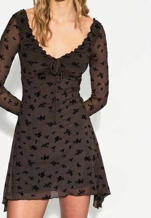 Kobieta ubrana w czarną, przezroczystą mini sukienkę z długim rękawem, ozdobioną małym kwiatowym wzorem i marszczonym wiązaniem z przodu.