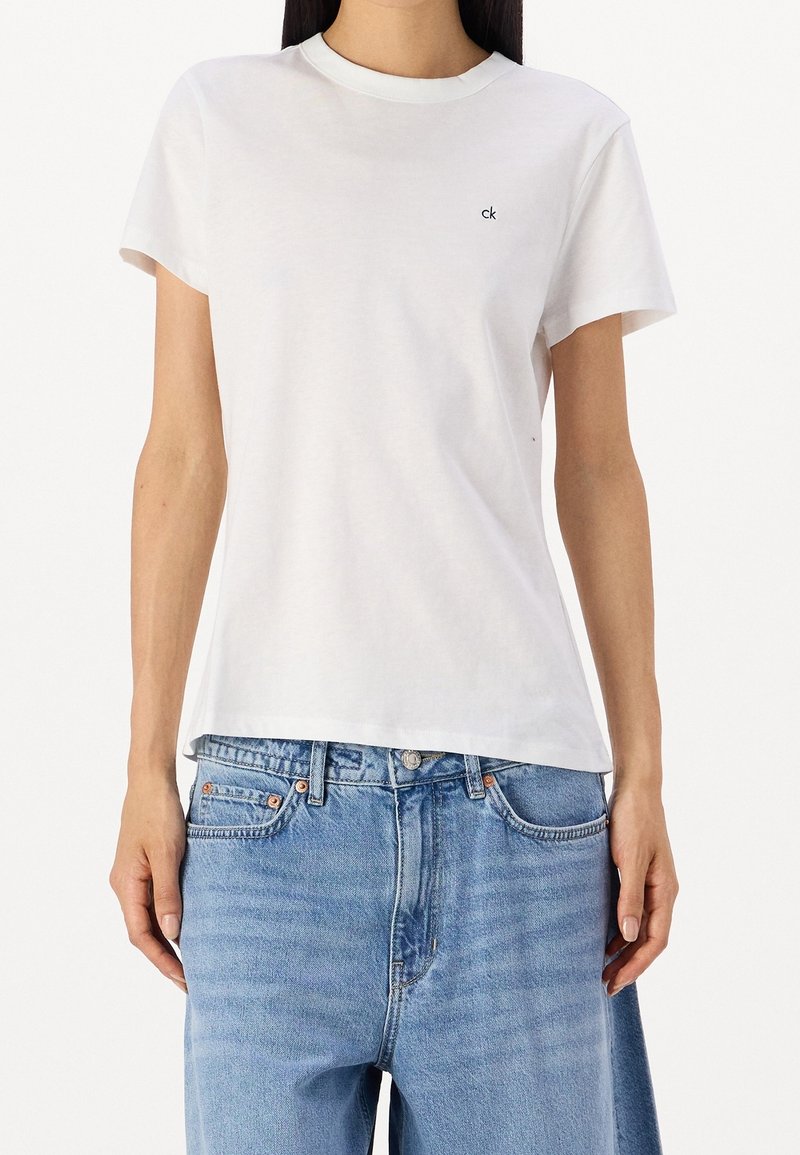 Femme portant un t-shirt blanc uni à manches courtes avec un petit logo "ck" sur la poitrine et un jean bleu ample.