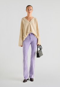 Beżowy sweter oversize z dekoltem w serek i prążkowanymi rękawami, połączony z jasnoliliowymi dzwonami. Akcesoria to mała czarna torebka.