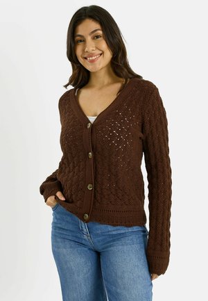 Mujer sonriente con cabello oscuro que lleva un cárdigan marrón de punto con botones sobre una camiseta blanca y jeans azules, de pie con una mano en el bolsillo.