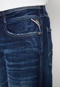 Dark blue denim jeans with a faded texture, contrast stitching, and a small logo tag in the front pocket. Standard five-pocket design.