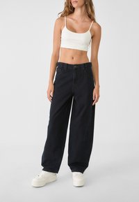 Jeans de mezclilla negros de pierna ancha con cintura alta, con bolsillos frontales y cierre de botón, combinados con un top corto blanco y zapatillas blancas.