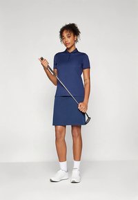 Mulher vestida com um conjunto de golfe azul-marinho, segurando um taco de golfe e posicionada contra um fundo cinza claro.