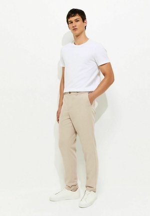 Jonge man staand met handen in de zakken, draagt een wit T-shirt, beige broek en witte sneakers tegen een effen witte achtergrond.