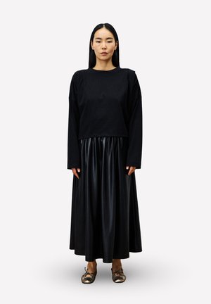 Femme avec de longs cheveux noirs portant un haut noir à manches longues, une jupe plissée noire brillante et des ballerines métallisées, se tenant devant un fond uni.