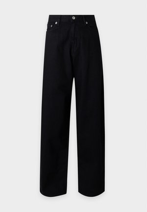 Mujer con abrigo marrón, suéter oscuro, pantalones negros, zapatos negros, sosteniendo un gran bolso de cuero marrón sobre un fondo claro liso.