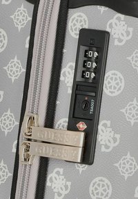 Superficie de maleta gris con patrones geométricos blancos, que presenta un candado combinado, un orificio para llave aprobado por la TSA y cremalleras plateadas grabadas con "GUESS".