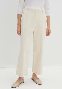 Cremfarbene Cordhosen mit weitem Bein und Gürtelschlaufen. Kombiniert mit hellbeigen, flachen, rutschfesten Schuhen. Glatte Textur und vertikale Streifen.