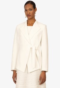 Veste blanche à encolure crantée, à manches longues et détail de ceinture à nouer. Tissu lisse, associée à une jupe texturée.