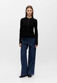 Svart ribbestrikket langermet polo med krage og knapper foran, kombinert med vide blå denimjeans og svarte sko.