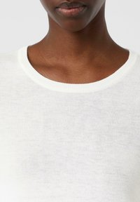 Primer plano del cuello y parte superior del pecho de una persona que lleva un suéter de punto blanco de cuello redondo frente a un fondo liso.