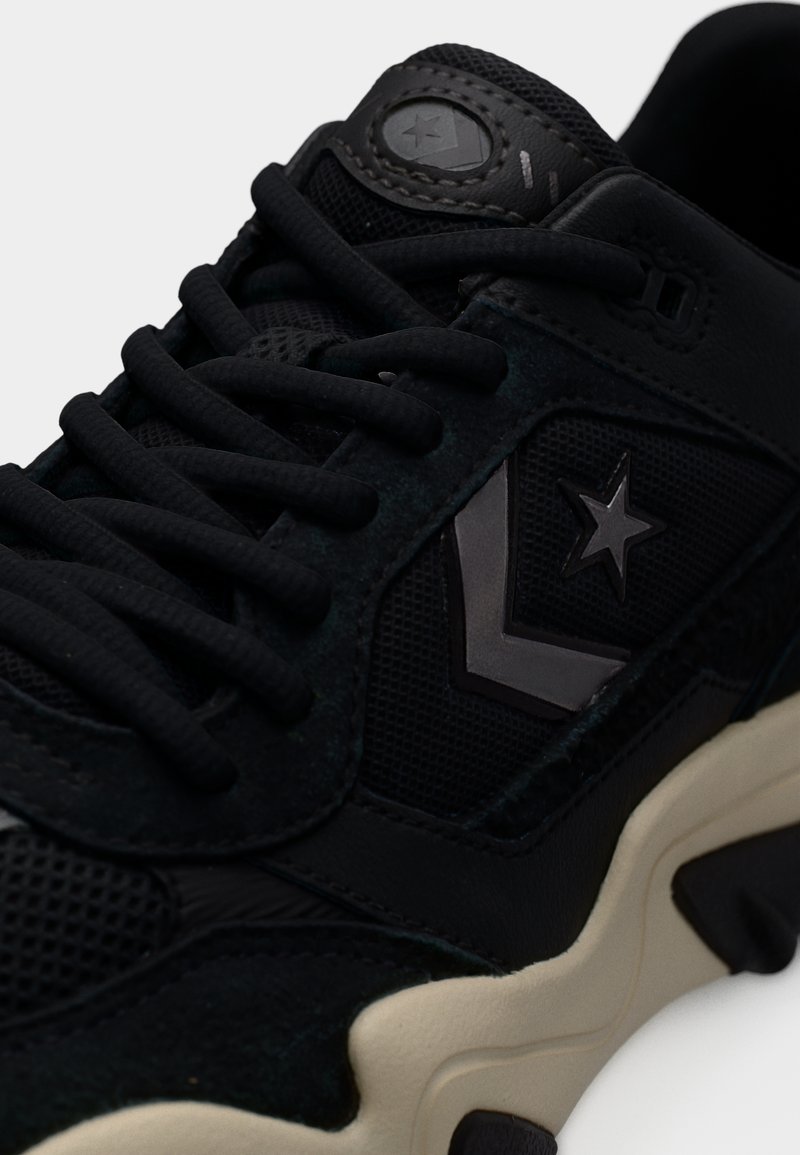 Zwarte sneaker met mesh- en suèdelagen, voorzien van dikke zwarte veters, logo-accenten en een dikke crèmekleurige zool.