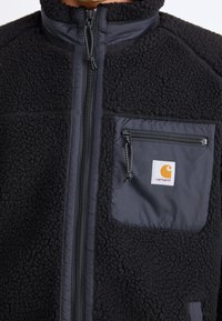 Sort fleecejakke med lynlås og en tekstureret overflade. Har en sort stofbrystlomme med lynlås og logo-patch.