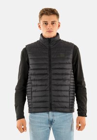 Zwarte gewatteerde bodywarmer met hoge kraag en ritssluiting. Horizontale stiksels en logopatch op de borst. Draagt men over een shirt met lange mouwen.