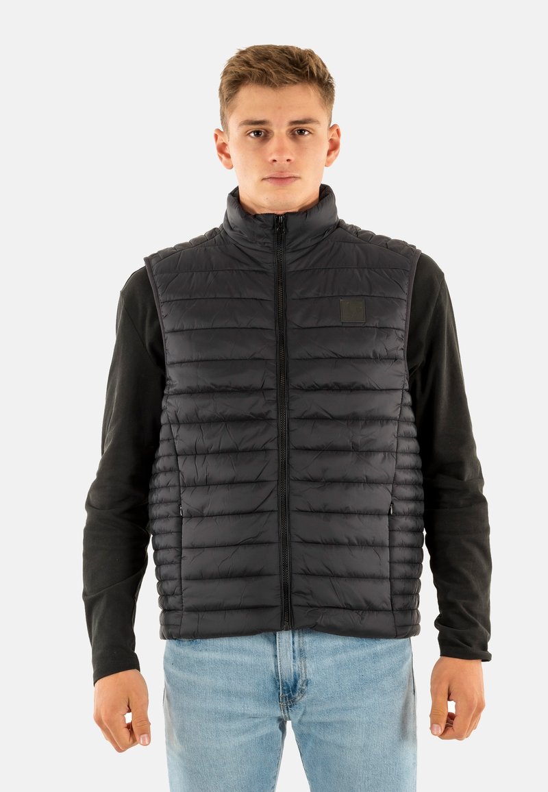 Zwarte gewatteerde bodywarmer met hoge kraag en ritssluiting. Horizontale stiksels en logopatch op de borst. Draagt men over een shirt met lange mouwen.