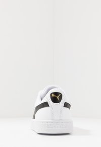 Baskets en cuir blanc avec accents noirs, semelle extérieure texturée et logo Puma doré à l'arrière. Forme arrondie et détail de col rembourré.