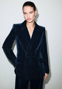 Mujer con el pelo hacia atrás, usando pendientes de aro de plata y un blazer de terciopelo azul oscuro de doble botonadura con pantalones a juego, frente a un fondo liso.