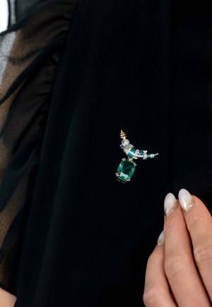 Una spilla in smeraldo e diamanti con un accento curvo in oro e pietre blu e trasparenti, attaccata a una superficie in tessuto nero.