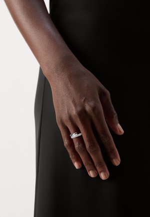 Silberner Ring mit mehreren dünnen Bändern, ausgestattet mit einem zentralen Stein und Verzierungen; getragen an einer Hand vor einem dunklen Hintergrund.