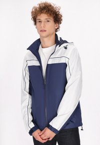 Marineblauwe en witte zip-up jas met capuchon, voorzien van elastische manchetten en een trekkoord. Lichtgewicht, gladde textuur en contrasterende kleurpanelen.