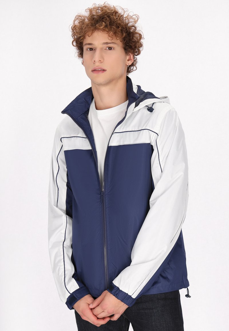 Marineblauwe en witte zip-up jas met capuchon, voorzien van elastische manchetten en een trekkoord. Lichtgewicht, gladde textuur en contrasterende kleurpanelen.