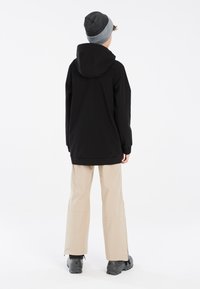 Schwarze Kapuzenjacke mit geripptem Saum, kombiniert mit beigen Hosen und schwarzem Schuhwerk. Graue Mütze fügt eine mehrschichtige Textur hinzu. Einfaches, funktionales Design.
