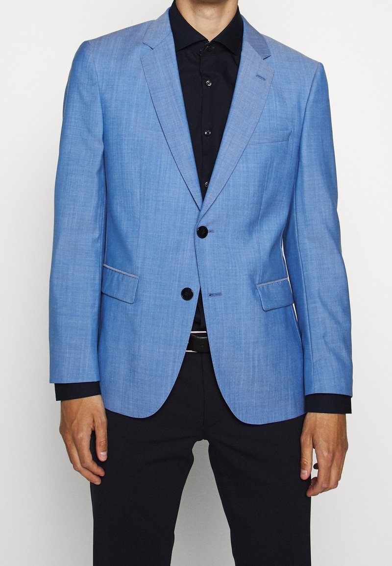 Blazer azzurro chiaro in un tessuto texturizzato. Presenta due tasche frontali, chiusura con un solo bottone e rever a punta. Indossato sopra una camicia nera.