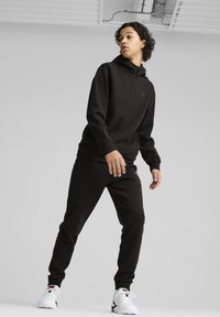 Joven de pie con una sudadera y pantalones negros de Puma y zapatillas blancas, mirando de lado contra un fondo claro y liso.