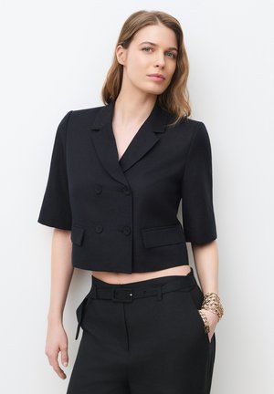 Mujer con blazer negro corto de doble botonadura y mangas hasta el codo, combinada con pantalones de talle alto a juego con cinturón.