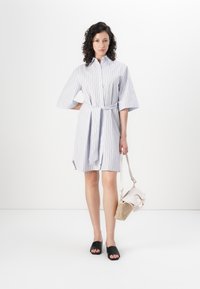 Vestido listrado com cintura ajustada por cinto, mangas curtas; feito de um tecido leve. Usado com sandálias pretas de slide e uma bolsa bege.