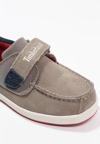 Zapato gris de ante tipo slip-on con una correa de velcro, suela blanca y forro rojo; presenta acentos azules y una marca bordada en la parte superior.