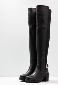 Botas negras por encima de la rodilla, hechas de cuero suave, con un tacón grueso, detalle de hebilla y suela texturizada.