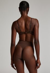 Braunes Lingerie-Set mit einem Bralette mit verstellbaren Trägern und einem passenden hochgeschnittenen Tanga. Glatte Textur und minimalistisches Design.