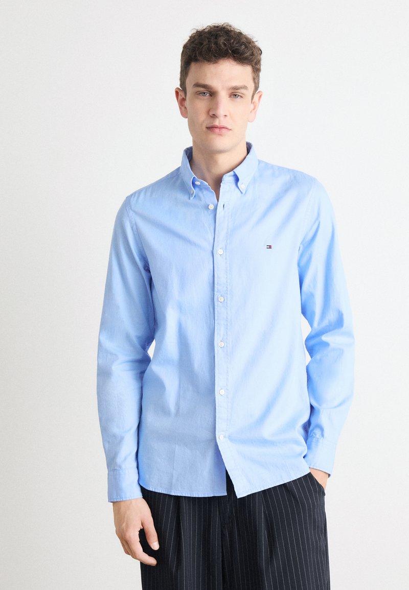 Camicia azzurra chiaro in cotone, con colletto classico e un piccolo logo sul petto. Indossata con pantaloni a righe.