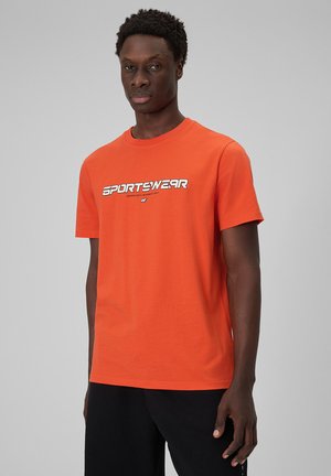 Mężczyzna w jasnopomarańczowej koszulce z krótkim rękawem z napisem „SPORTSWEAR” i czarnych spodniach, stojący na jednolitym szarym tle.
